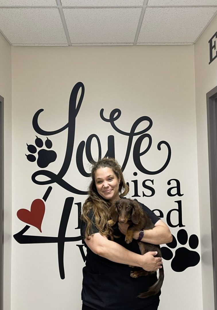 A woman holding her dog in front of a wall with words.